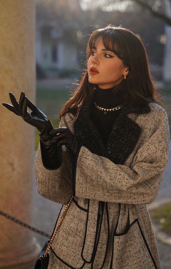 Woman in stylish winter coat and black gloves, standing outdoors, looking curious, with a golden chain purse.