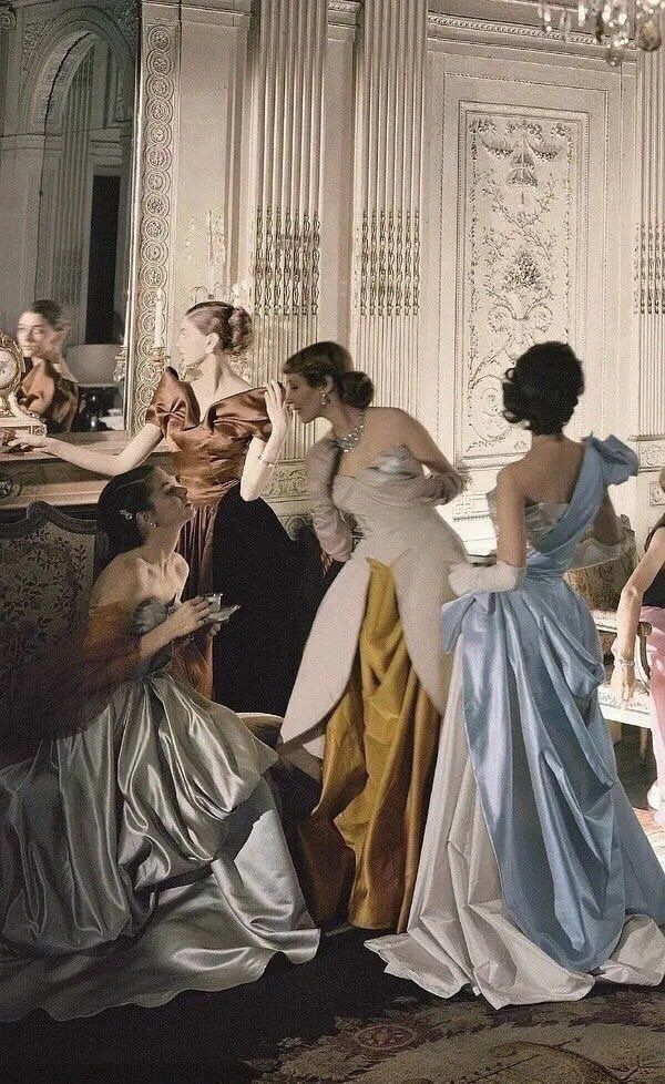 Elegant women in vintage gowns socializing in an opulent, ornately decorated room.