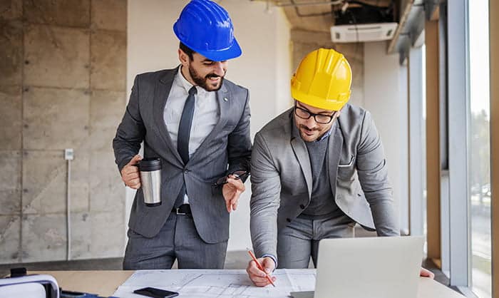 Two architects in suits and hard hats discussing blueprints at a desk with a laptop in an office setting.