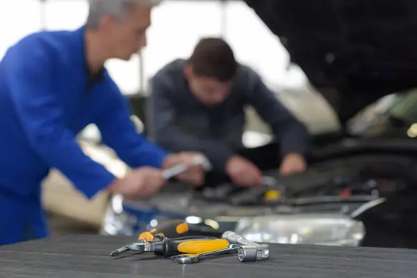 Mechanics working on car engine, focus on tools in foreground, in auto repair shop.