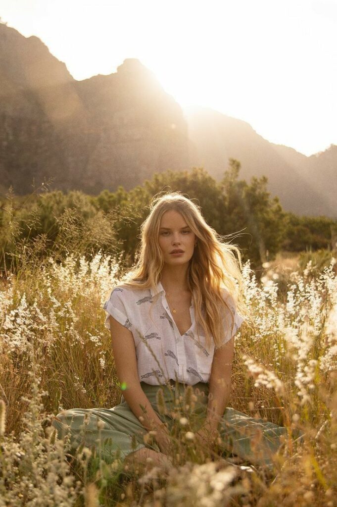 Woman in a meadow during sunset, surrounded by grass and mountains, wearing a white blouse and green skirt.