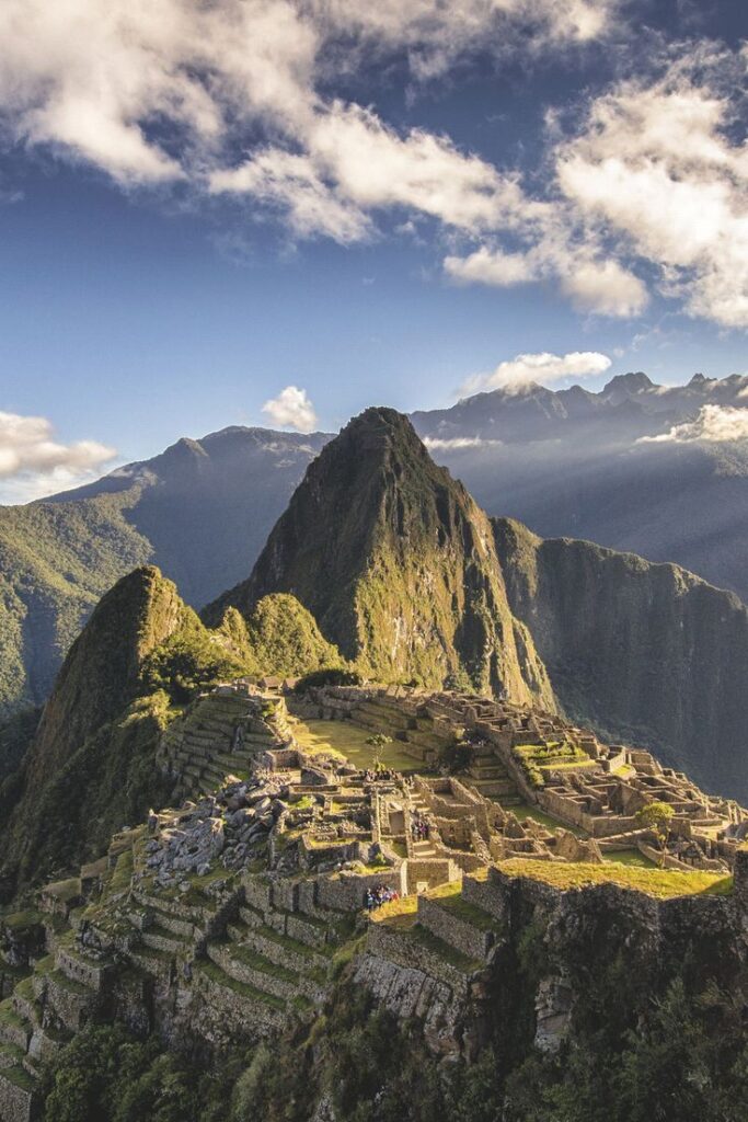 Machu Picchu ruins with Huayna Picchu mountain in background under a cloudy sky, showcasing lush green terraces.