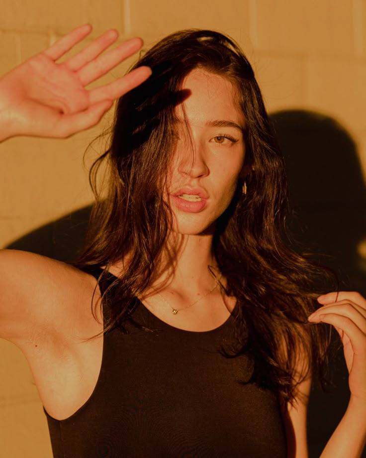 Woman shielding her eyes from the sun, wearing a black top, with hair flowing, against a beige wall background.