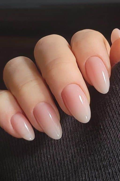 Elegant almond-shaped nails with nude polish, close-up view, against a dark fabric backdrop.