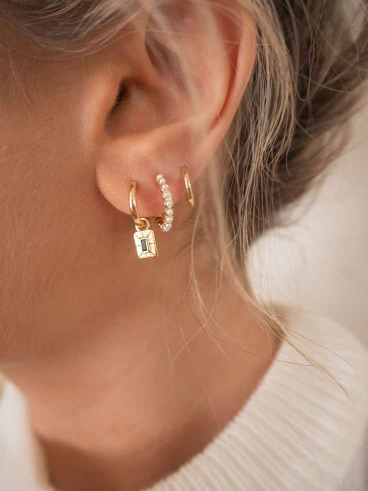 Close-up of a woman's ear wearing three dainty gold hoop earrings, one with pearls and a gold pendant.