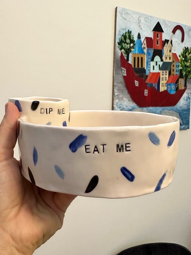 Hand holding a ceramic bowl with the text DIP ME EAT ME and a colorful painting in the background.