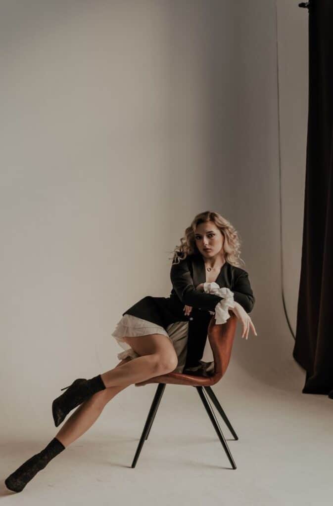 Fashion model poses in stylish outfit on chair, minimal studio backdrop.