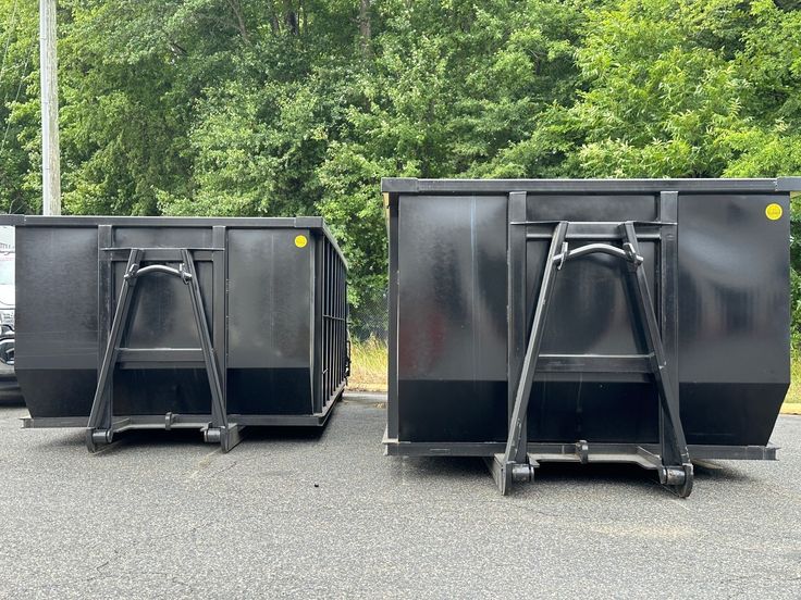 Two black dumpsters on pavement with green trees in background, ideal for waste management or recycling solutions.