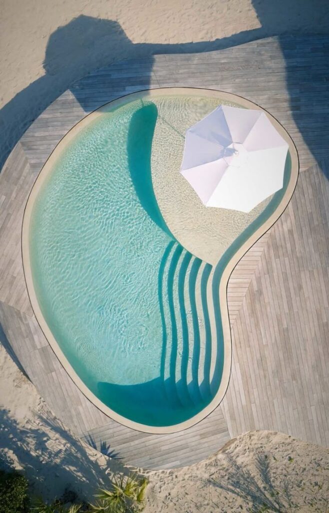 Aerial view of a kidney-shaped pool with clear water, wooden deck, and white umbrella on a sandy beach setting.