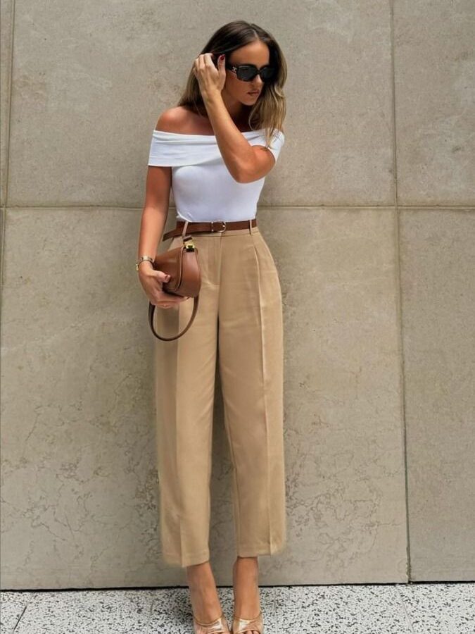 Stylish woman in beige pants, white top, sunglasses, holding a brown purse, standing against a concrete wall.