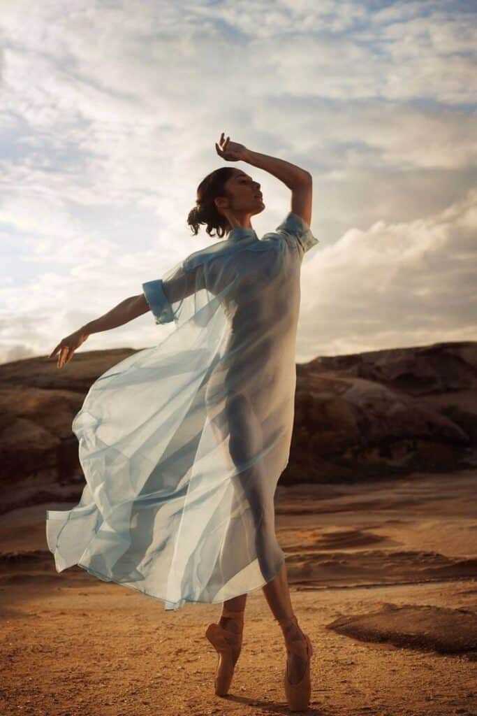 Ballet dancer in flowing blue dress gracefully poses on a sandy landscape under a cloudy sky.