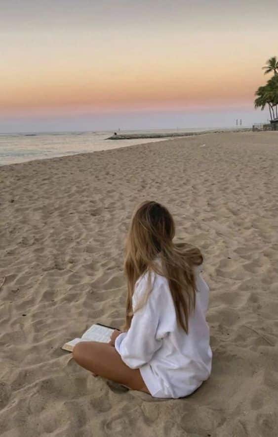 Woman in a white sweater reading on a beach at sunset, with palm trees in the distance.