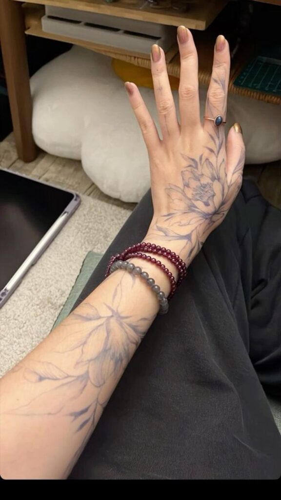 Hand with floral tattoos, wearing bracelets and a blue stone ring, resting on dark fabric near a tablet and cushion.