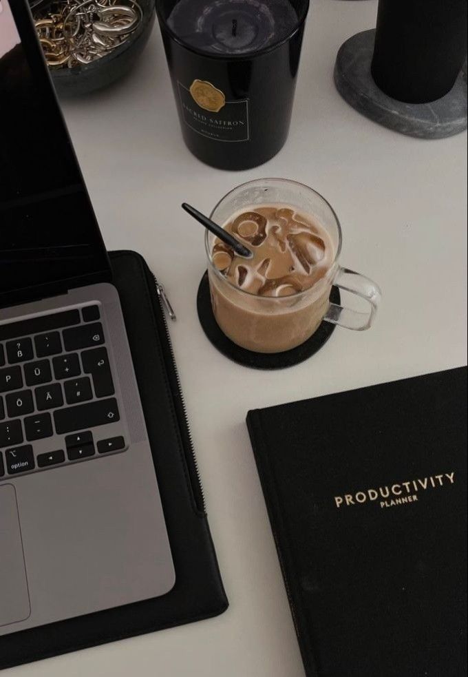 A workspace with a laptop, iced coffee, candle, and productivity planner on a desk for effective work environment setup.