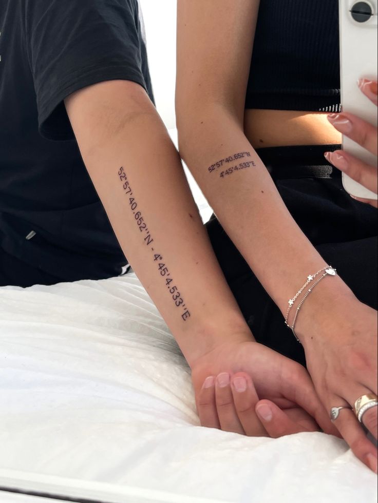 Two forearms with matching longitude and latitude coordinate tattoos, one in a black shirt and one in a black crop top.