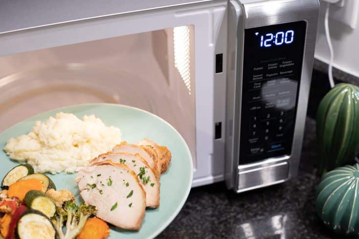 Plate of turkey, vegetables, and mashed potatoes in front of a microwave, digital clock showing 12:00.