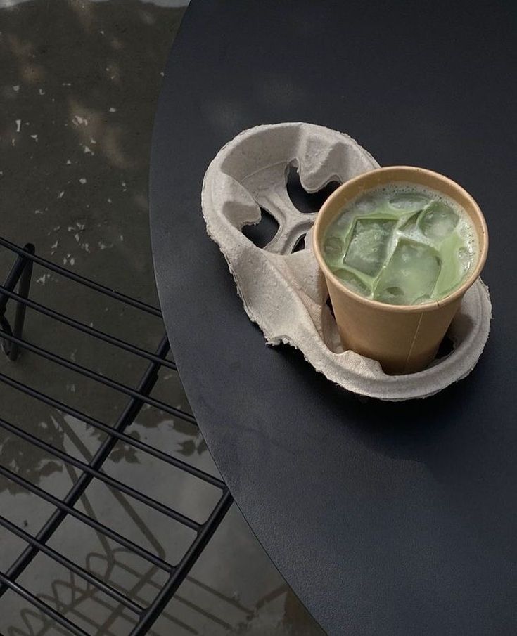 Iced matcha latte in a paper cup holder on a sleek black metal table, with reflections on a wet surface.