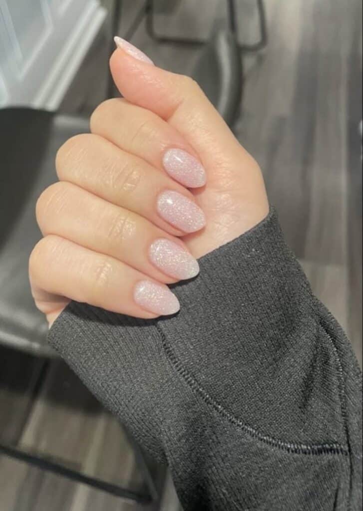 Close-up of a hand with sparkly pink almond-shaped nails, wearing a dark sleeve, on a wooden floor background.
