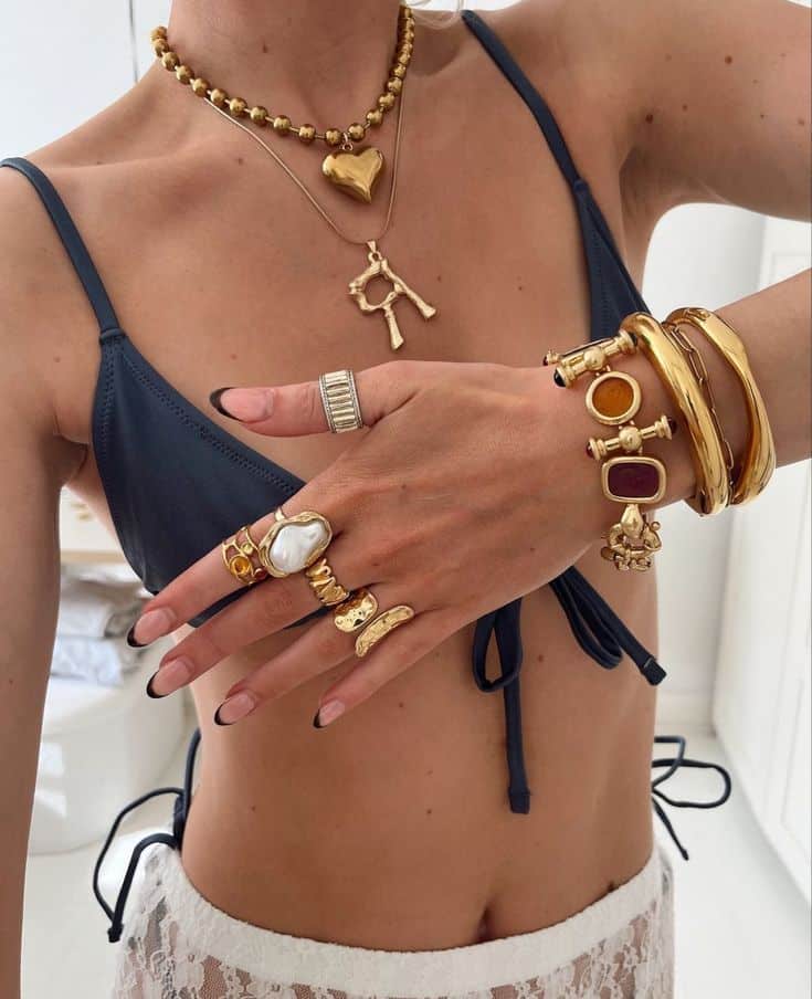 Woman wearing gold jewelry, including layered necklaces, rings, bracelets, and a bikini top. Fashion accessories.