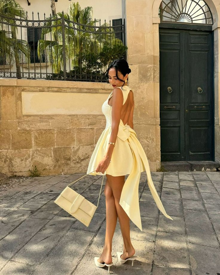 Woman in elegant yellow dress with bow walks, holding handbag in front of vintage door, sunny day.