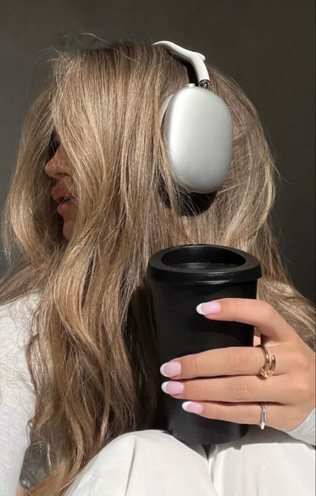 Woman with long blonde hair, wearing headphones, holds a black cup. Pink and white manicure highlighted in sunlight.