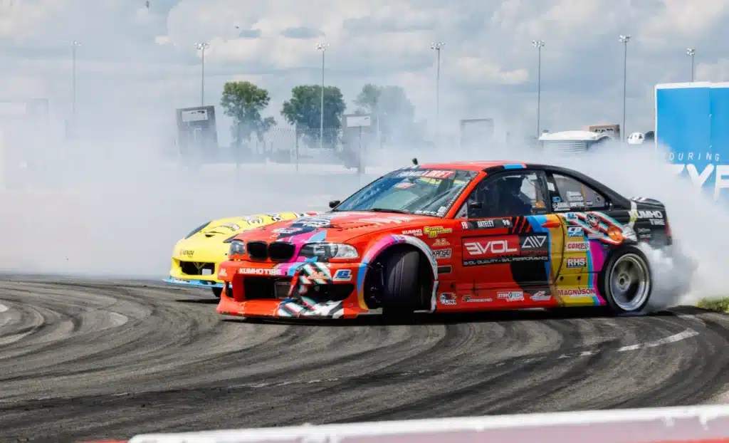 Colorful cars performing drift maneuvers on a race track, creating smoke against a clear sky.