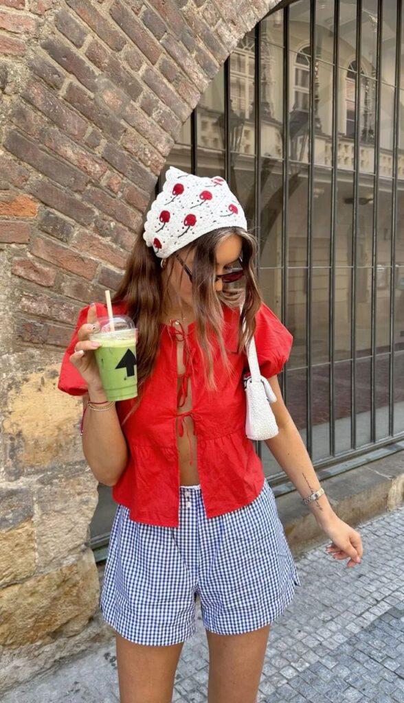 Trendy woman in a red top, gingham shorts, and mushroom hat, holding a green smoothie outdoors. Fashionable summer look.