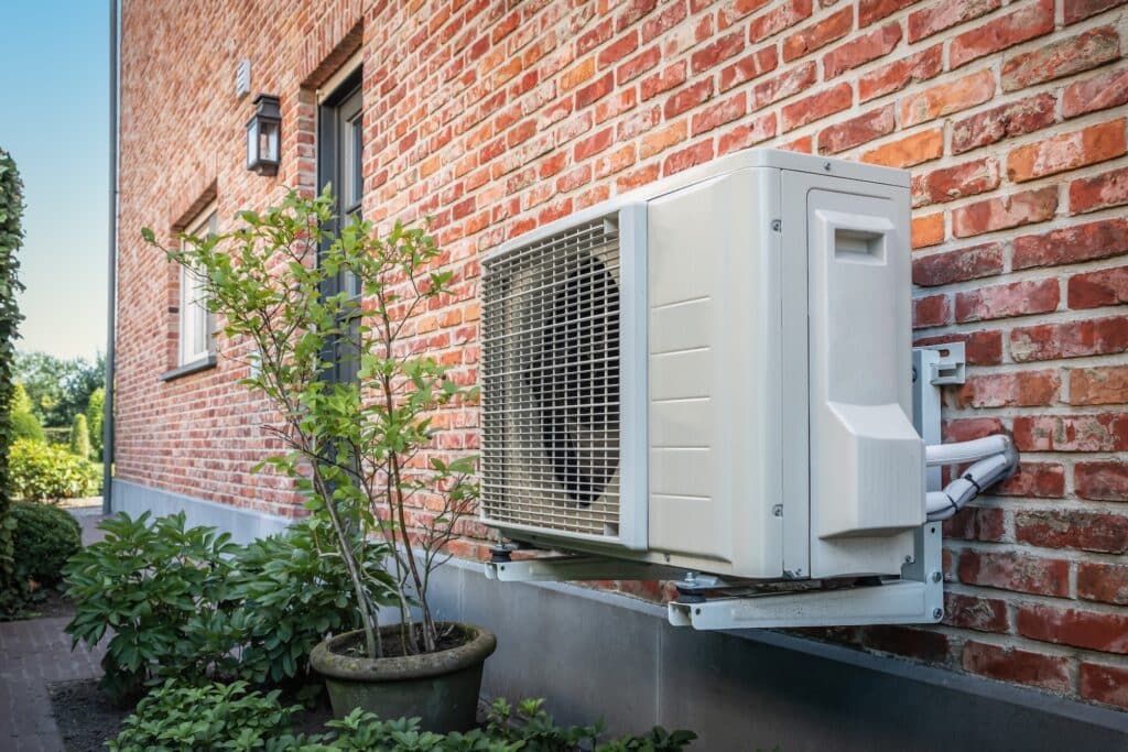 Outdoor air conditioning unit on a brick wall with nearby plants, providing energy-efficient cooling.