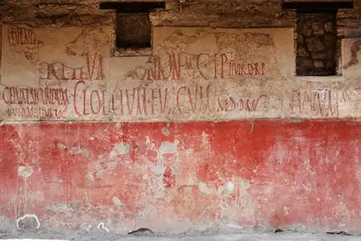 Ancient Roman graffiti on a weathered red and beige wall, showcasing historical inscriptions and art in a Pompeii archaeological site.