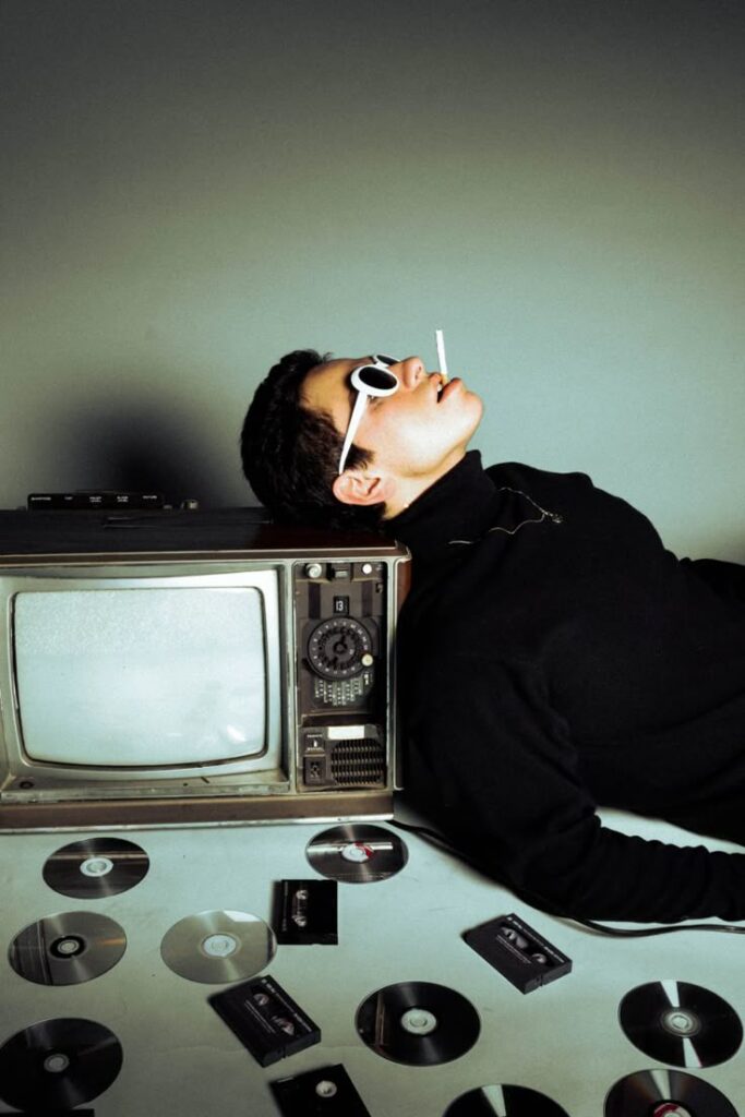 Retro-themed scene with a person in sunglasses leaning on a vintage TV, surrounded by CDs and VHS tapes.