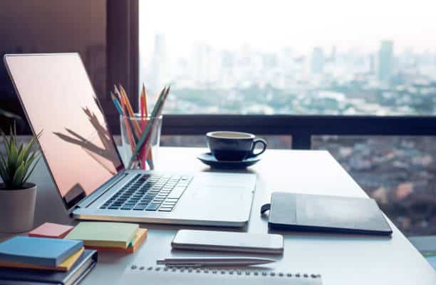 Modern workspace with laptop, coffee cup, and stationery overlooking cityscape through large window.