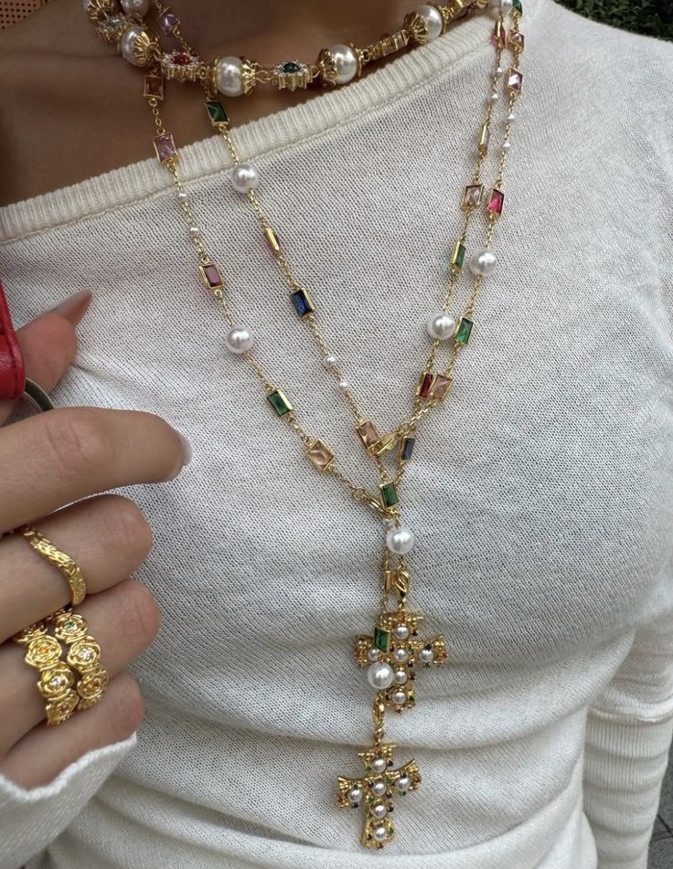 Layered gold necklaces with pearls and colorful gems on white sweater, featuring cross pendants and matching rings.