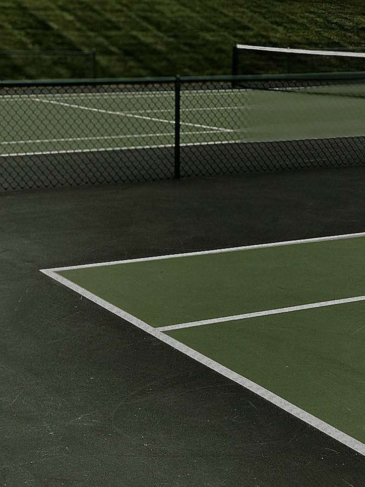 Empty tennis court with green surface, surrounded by a fence, under cloudy skies. Ideal for a peaceful game.