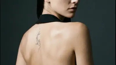 Elegant woman in black backless dress looking over shoulder, small koi fish tattoo on upper back, minimalist studio portrait