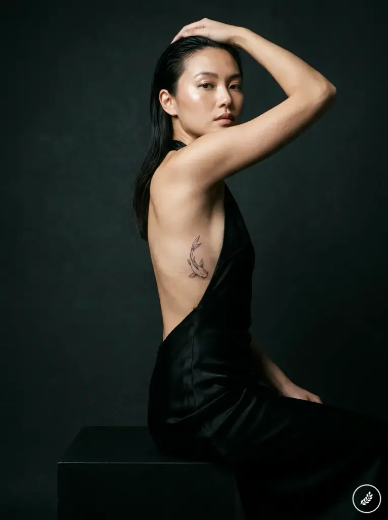 Studio portrait of woman in backless black dress showing delicate koi fish rib tattoo, elegant side pose on dark background