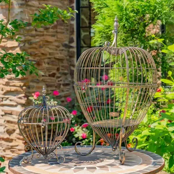 lesera-2 Decorative iron birdcages on a patio table in a lush garden with pink flowers and stone wall background. | Sky Rye Design Decorative iron birdcages on a patio table in a lush garden with pink flowers and stone wall background.