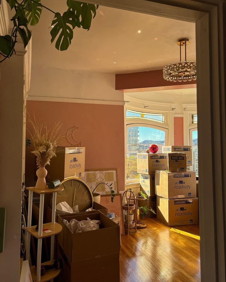 Room filled with moving boxes, sunlight streaming in, highlighting the wooden floor and modern decor elements.