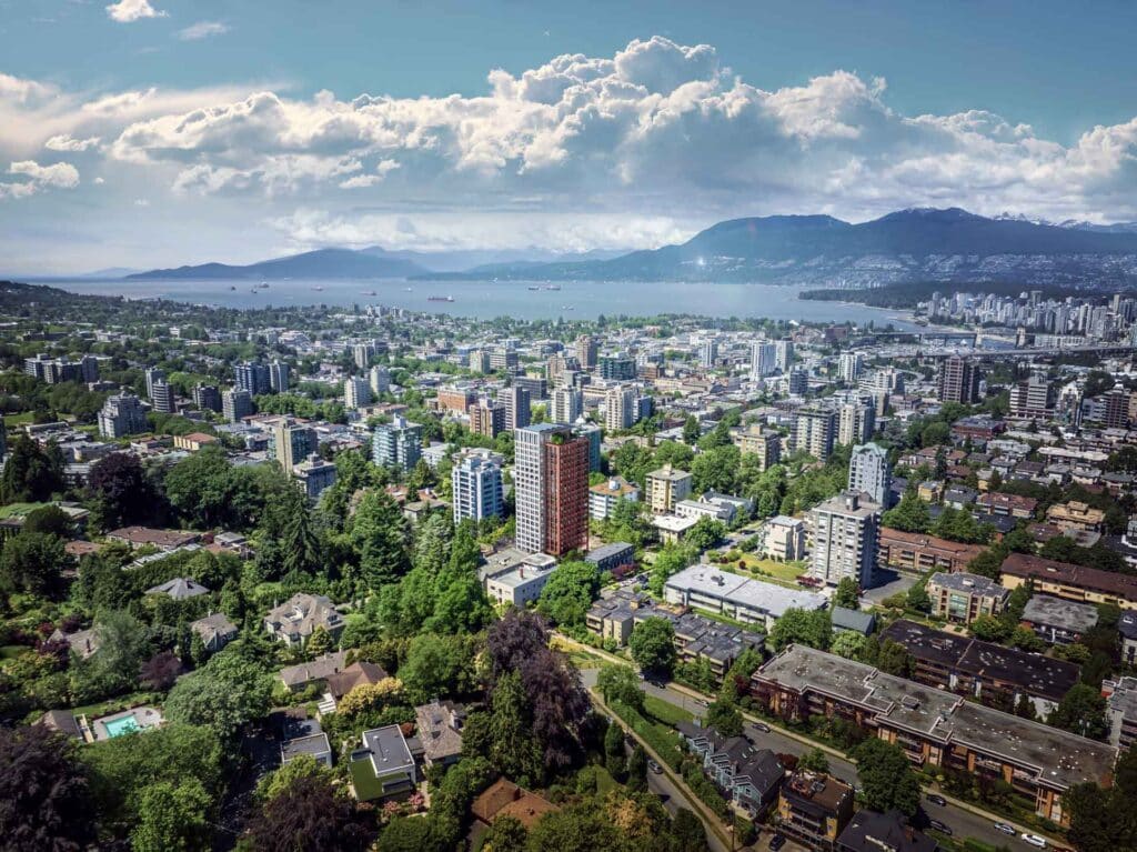 Aerial view of Vancouver cityscape with lush greenery, tall buildings, and distant mountains under a cloudy sky.