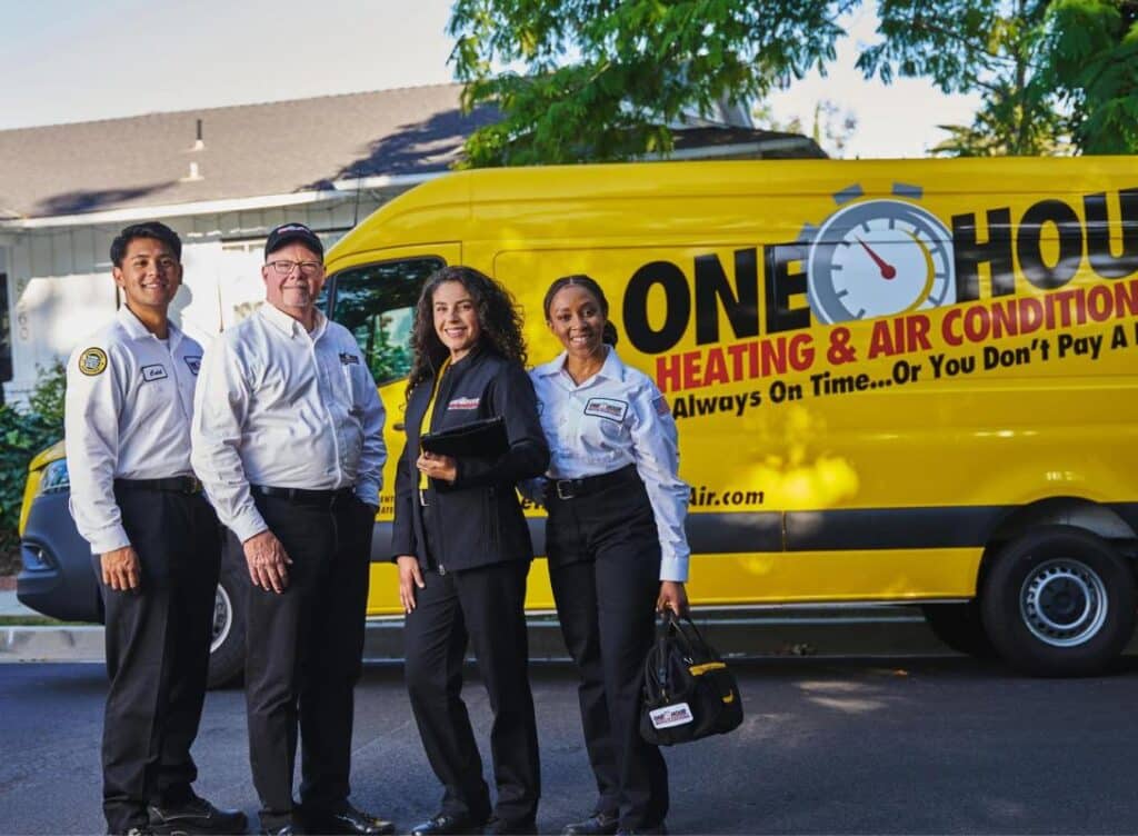 Four HVAC technicians smiling in front of a yellow service van from One Hour Heating & Air Conditioning.