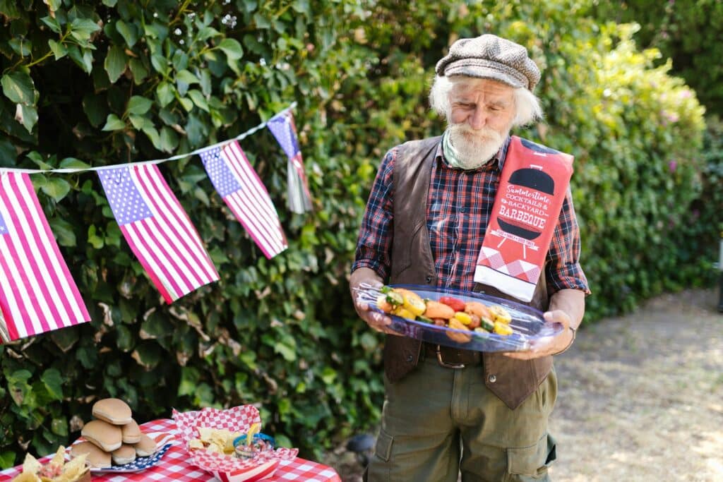 pexels-photo-8523490 Elderly man at summer barbecue holding a tray of grilled vegetables, with American flags and a checkered table setup. | Sky Rye Design Elderly man at summer barbecue holding a tray of grilled vegetables, with American flags and a checkered table setup.