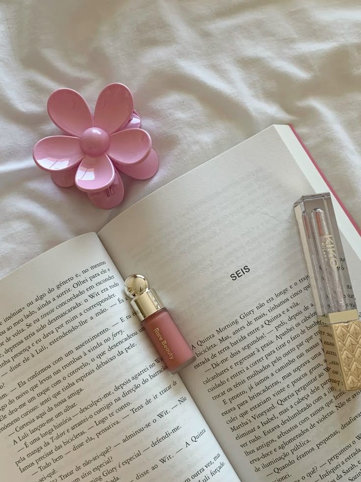 Open book on a bed with a pink flower hair clip and lip glosses nearby, creating a cozy reading vibe.