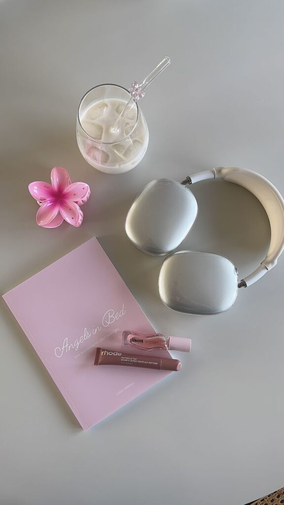 Silver headphones, pink book, iced drink, and lip gloss on a table with a pink flower. Calm and relaxing setup.