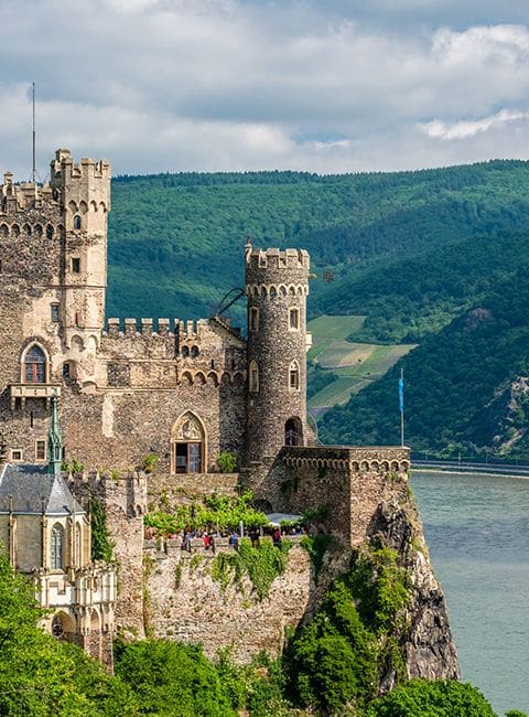Historic castle perched on a lush cliff by a river, surrounded by scenic green hills under a cloudy sky.
