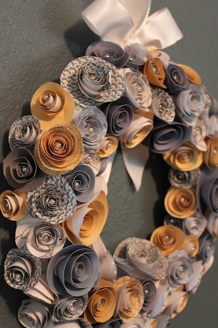 Handmade wreath with rolled paper flowers in gray and yellow tones, tied with a white ribbon on a green wall.