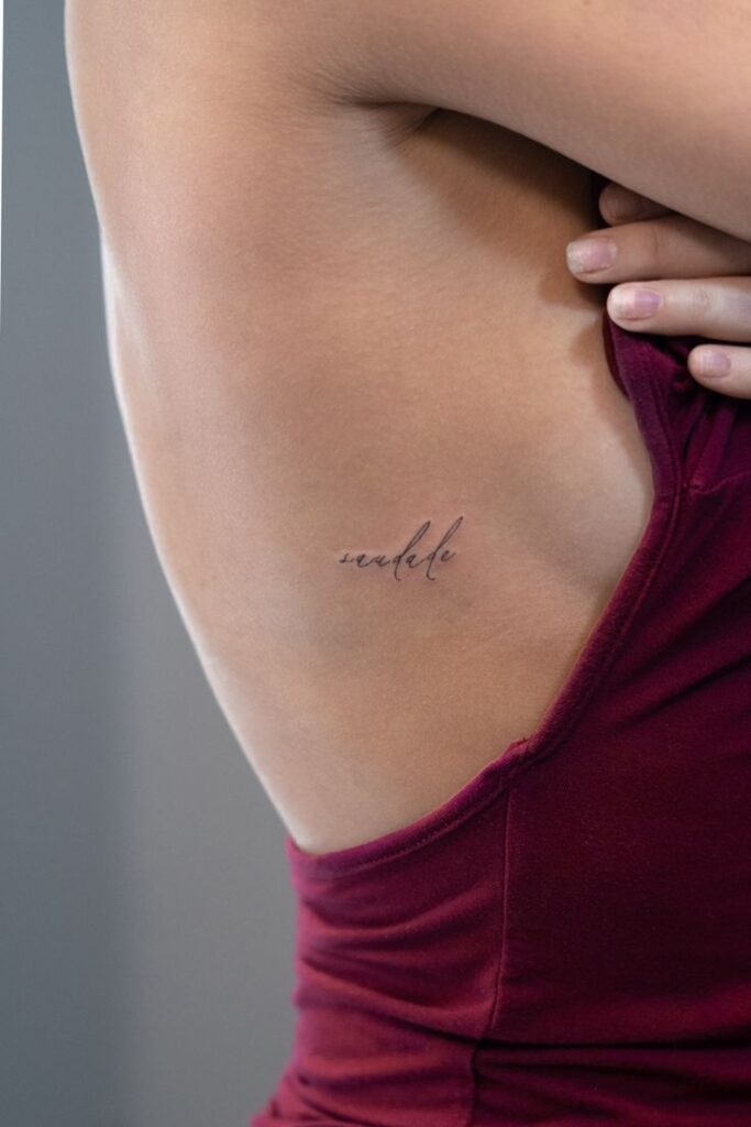 Elegant script tattoo on woman's side under maroon top, showcasing minimalistic body art style.
