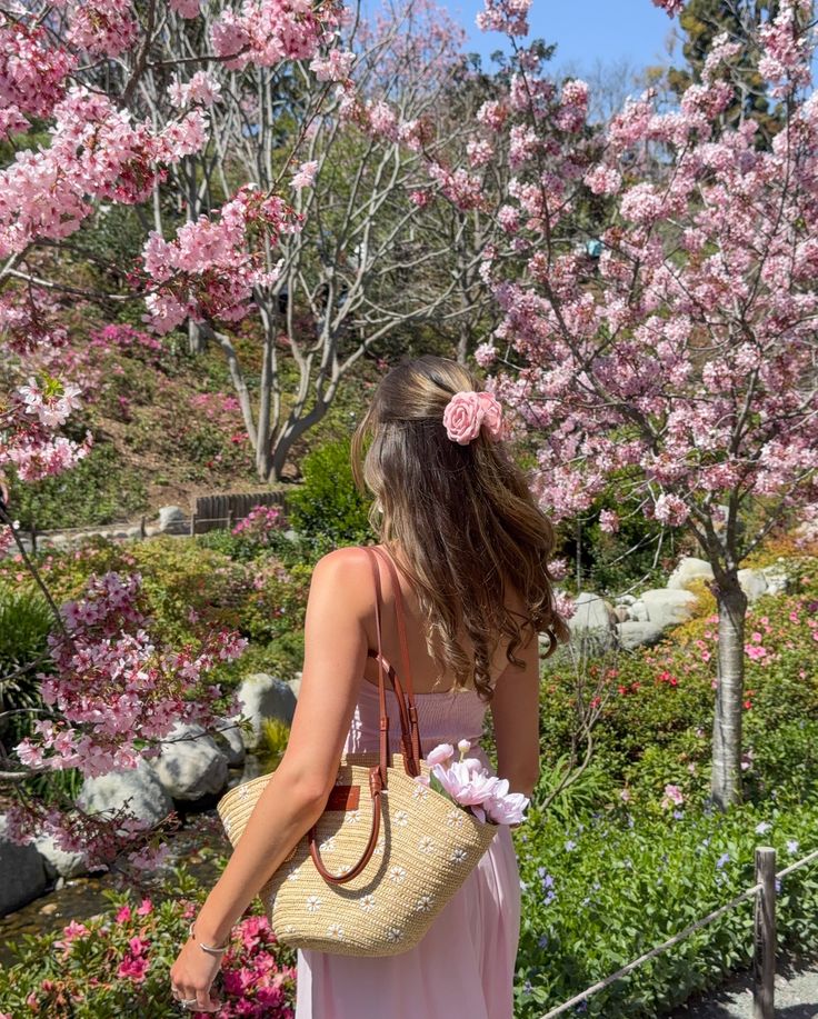 spring is springing 🌸🌼🌺🌷☀️ Woman in pink dress with floral hairpin and straw bag admires cherry blossoms in a sunlit garden. | Sky Rye Design Woman in pink dress with floral hairpin and straw bag admires cherry blossoms in a sunlit garden.