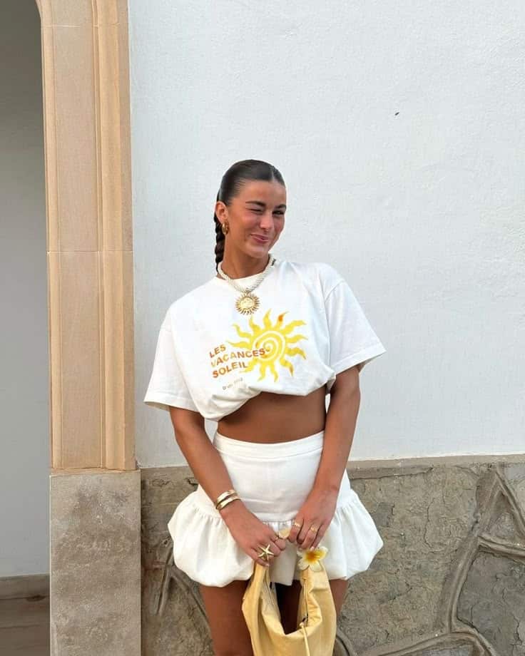 Woman in stylish white outfit with sun print shirt, winking and holding a yellow bag in front of a stone wall.