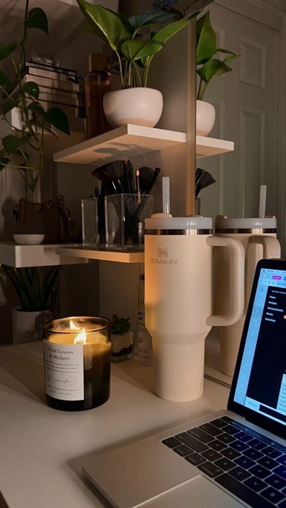 Cozy desk with candle, laptop, and large cup; decorated with plants and organized shelves.