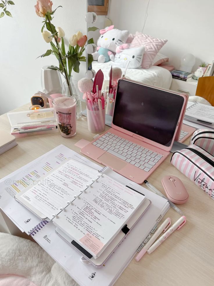 Cozy pink-themed study desk setup with laptop, notes, pens, and flowers for productive work or study session.