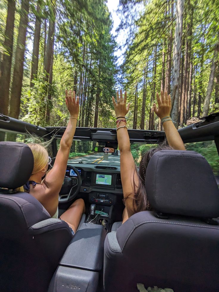 Two people enjoying a forest drive with hands raised in a convertible, surrounded by tall trees.